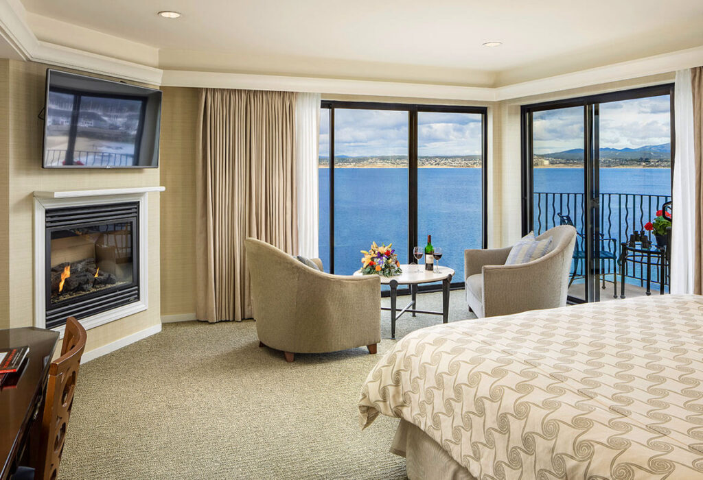 Upscale guest bedroom with bed arm chairs fireplace leading to huge windows overlooking the Pacific Ocean hotels in Monterey CA