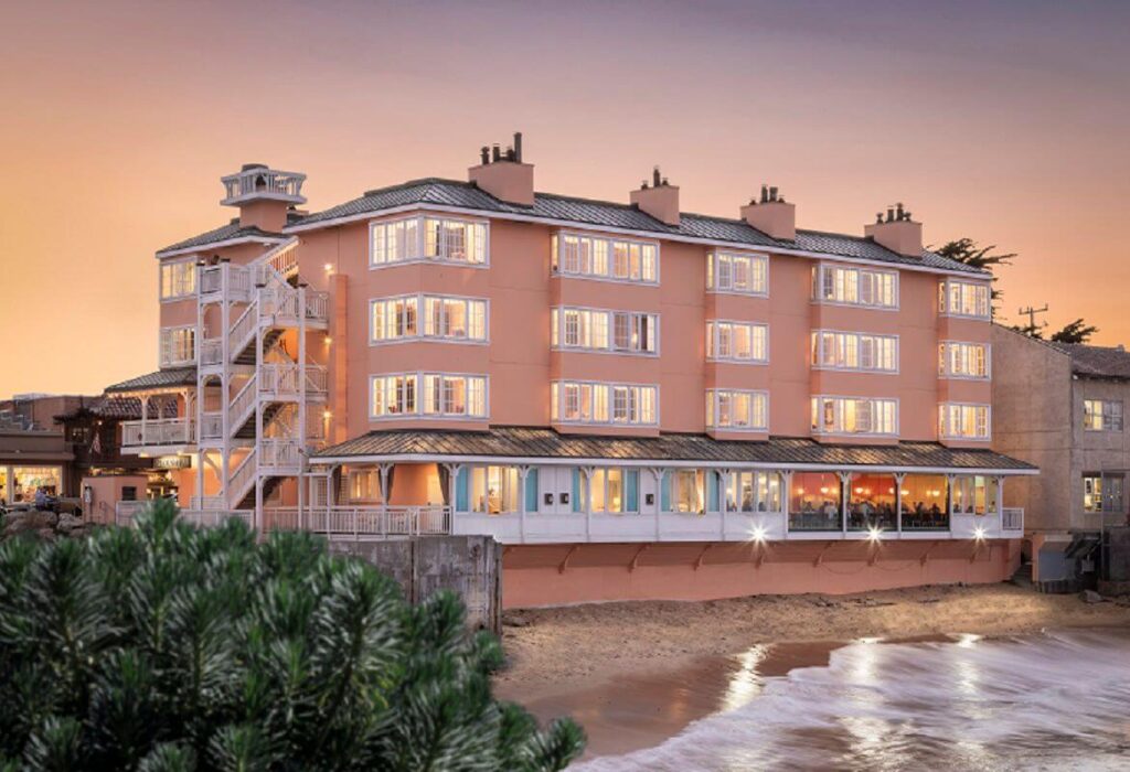 Exterior photo of a four or five story long building colored a soft pink or salmon at sunset with waves lapping onto a beach at the entrance one of the top affordable hotels in Monterey California