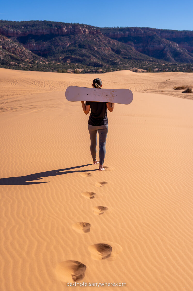 Person walking on sand dune with sandboard on back and leaving fresh footprints