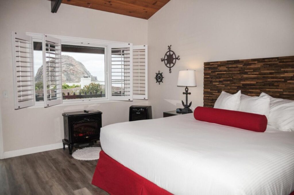Guest bed with red and white decor on a wooden floor with small fire place on ground and open window