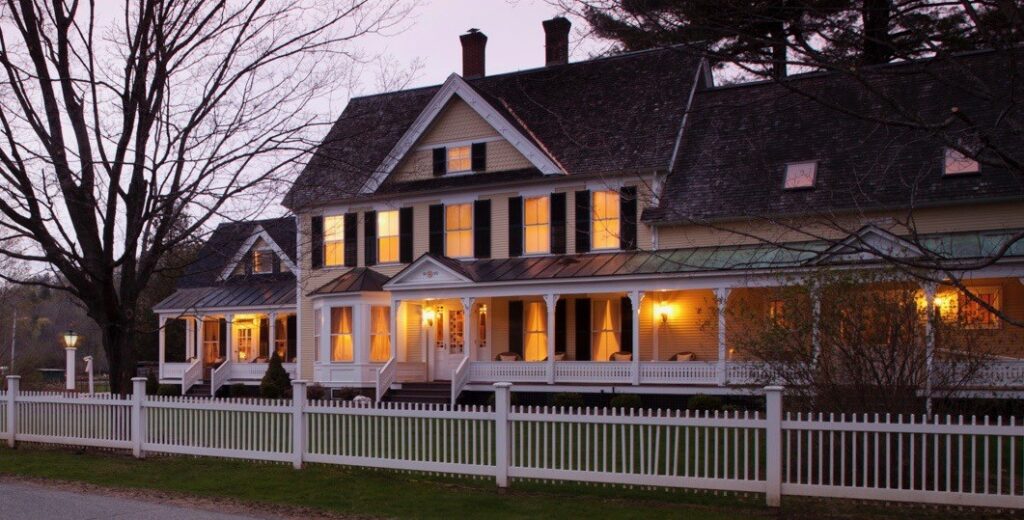 Exterior photo of a bed and breakfast taken at dusk with lights in windows and a white fence around the house