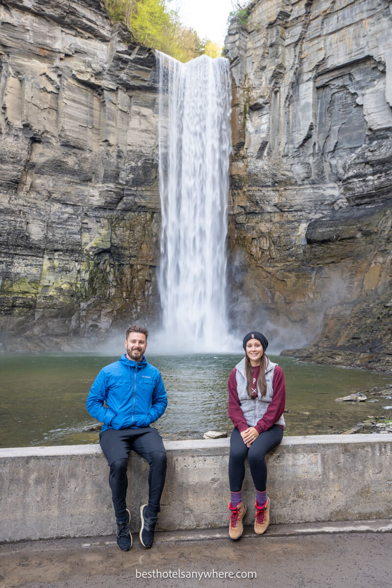 Couple sat on a small wall either side of a tall and narrow waterfall in the background