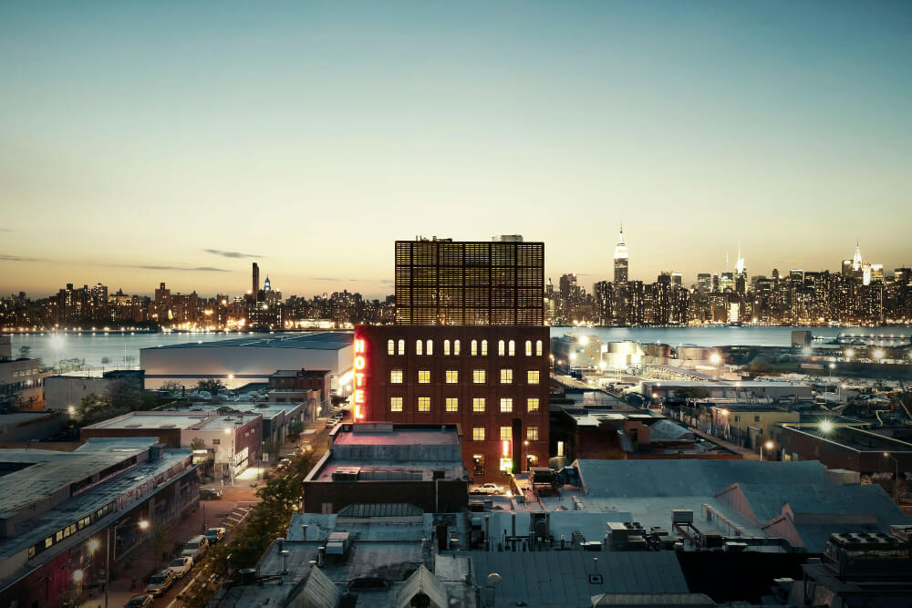 Wythe Hotel in Williamsburg NY lit up at night with Manhattan skyline in background