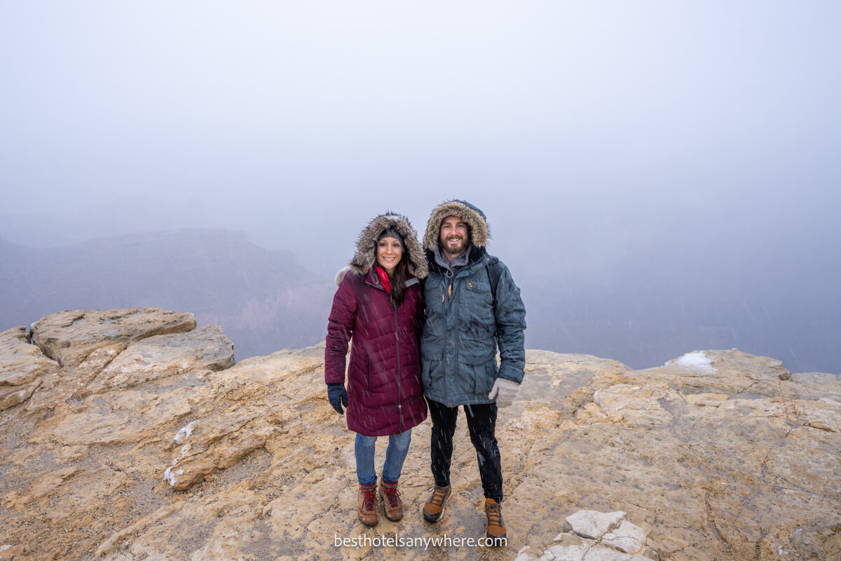 Couple standing together in winter coats on the rim of Grand Canyon in light snow and heavy clouds