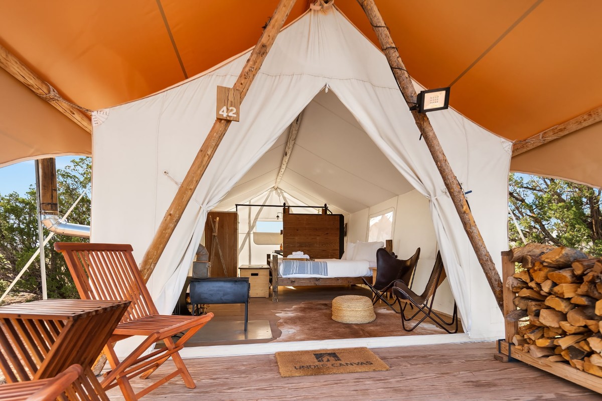 Luxury glamping tent with bed, table and chairs inside, and a pile of firewood under awning outside