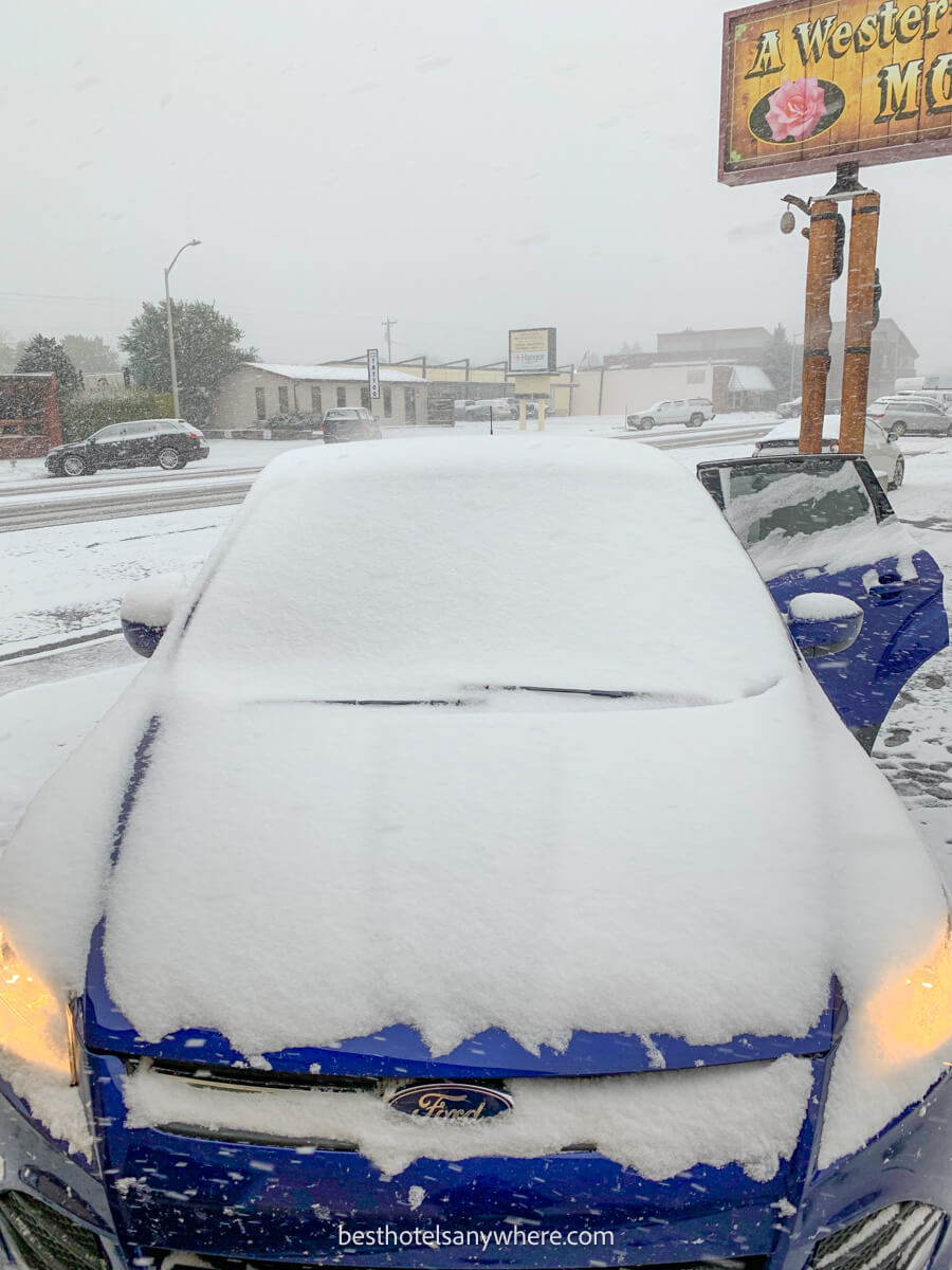 Blue car with headlights on covered in snow and snow falling heavily near a motel in Cody WY