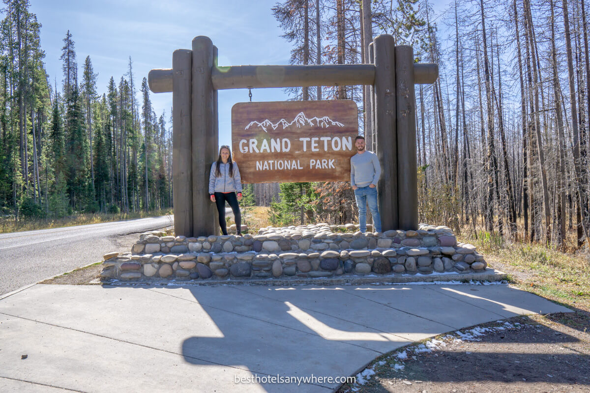 Couple standing on either side of the Grand Teton welcome sign on a sunny day