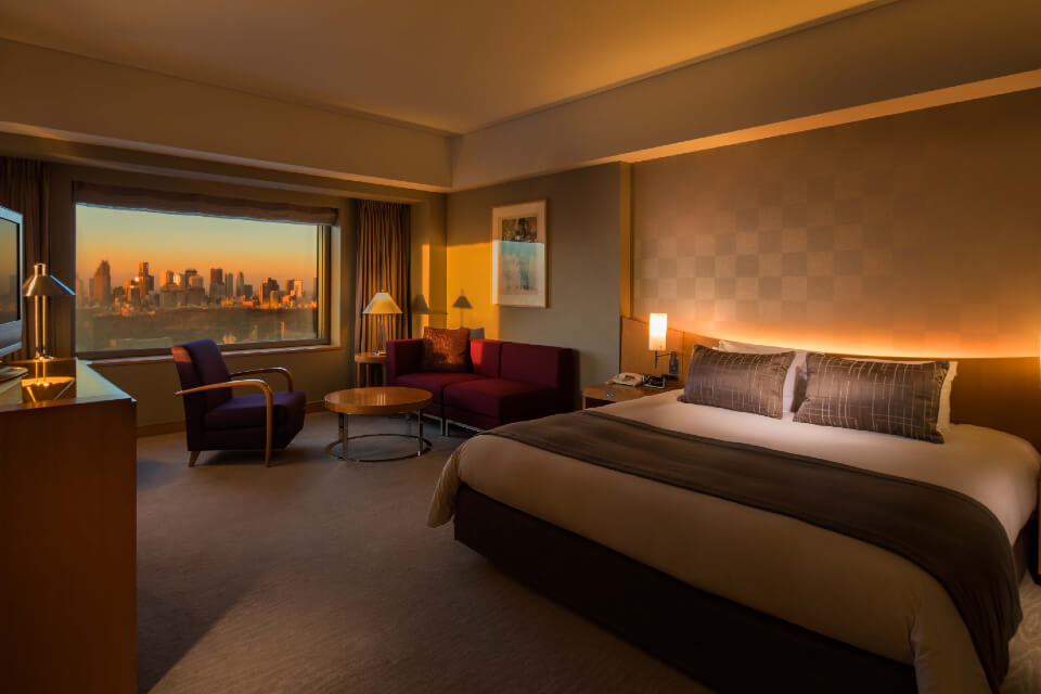 Inside a dimly lit Tokyo hotel guest room with bed and seating area leading to window with a city view