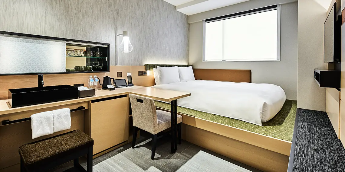 Inside a small guest bedroom at a cheap Tokyo hotel with bed on wooden platform and desk with small window