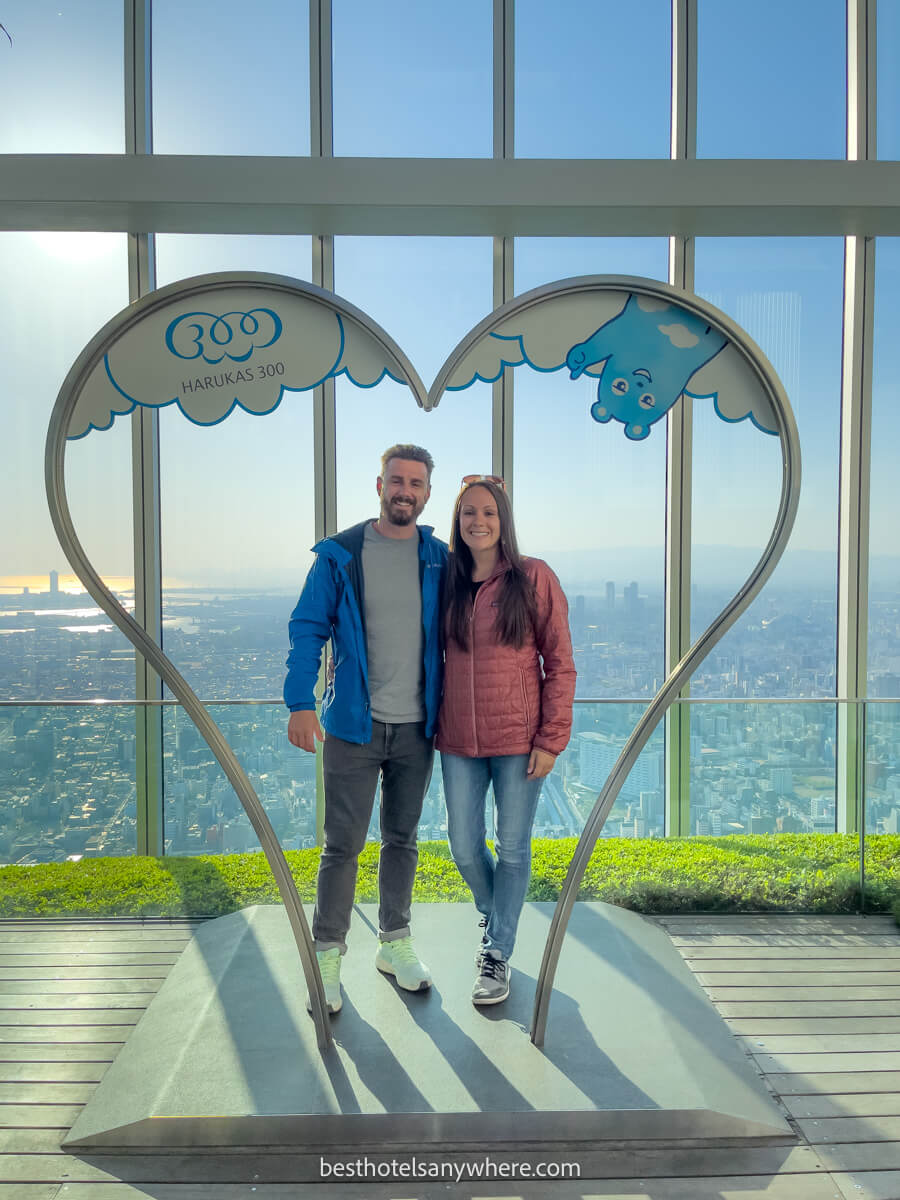 couple standing together for a photo inside a heart shaped metal frame in late afternoon with tall glass windows behind overlooking Osaka