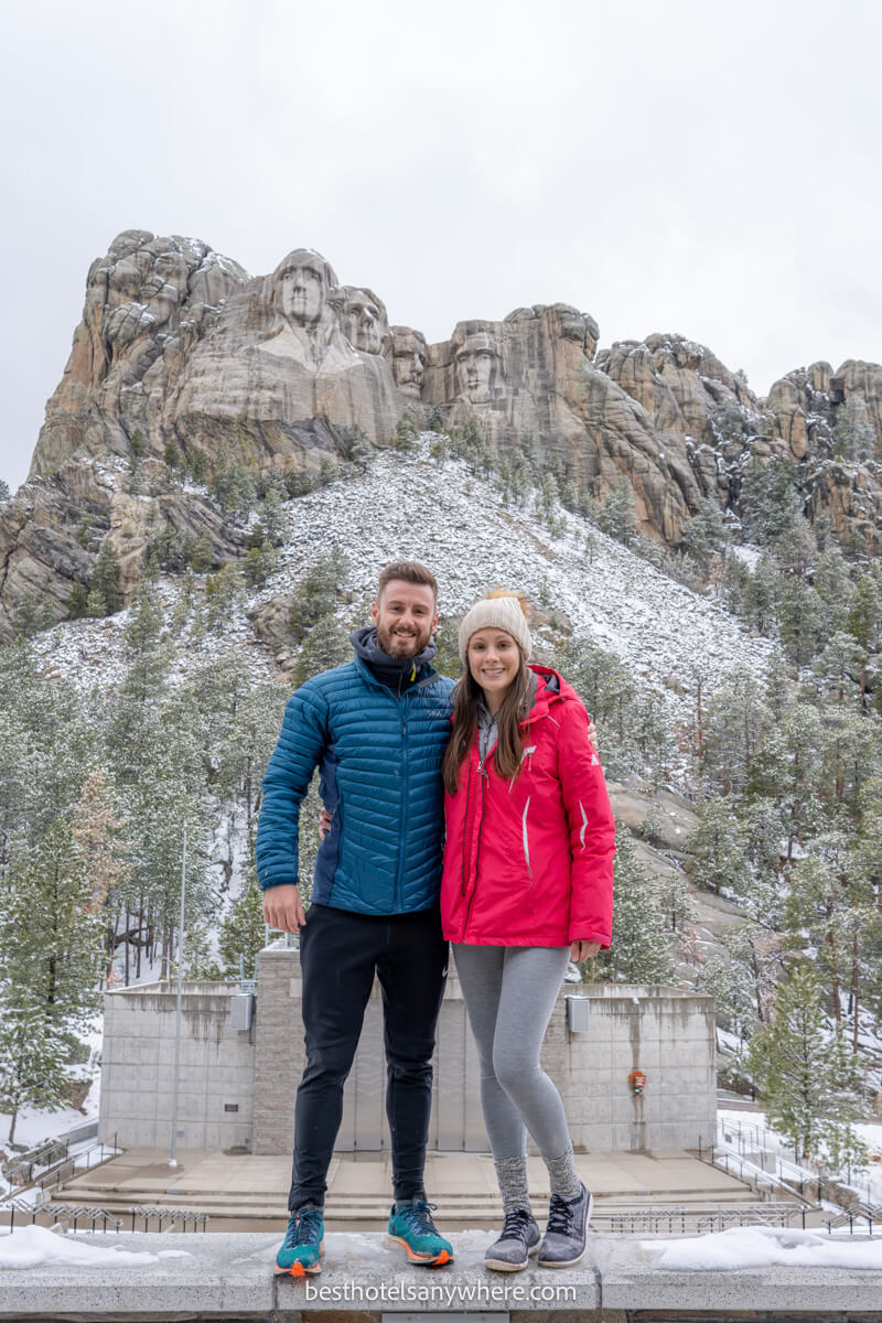 Couple standing together for a photo in front of Mount Rushmore with snow on the ground