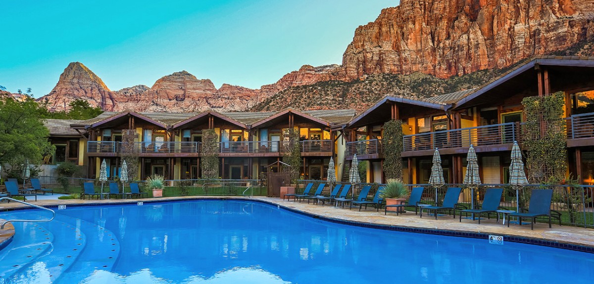 Photo of a blue outdoor swimming pool with hotel rooms around the edges and red rock cliffs behind at a Springdale Utah hotel