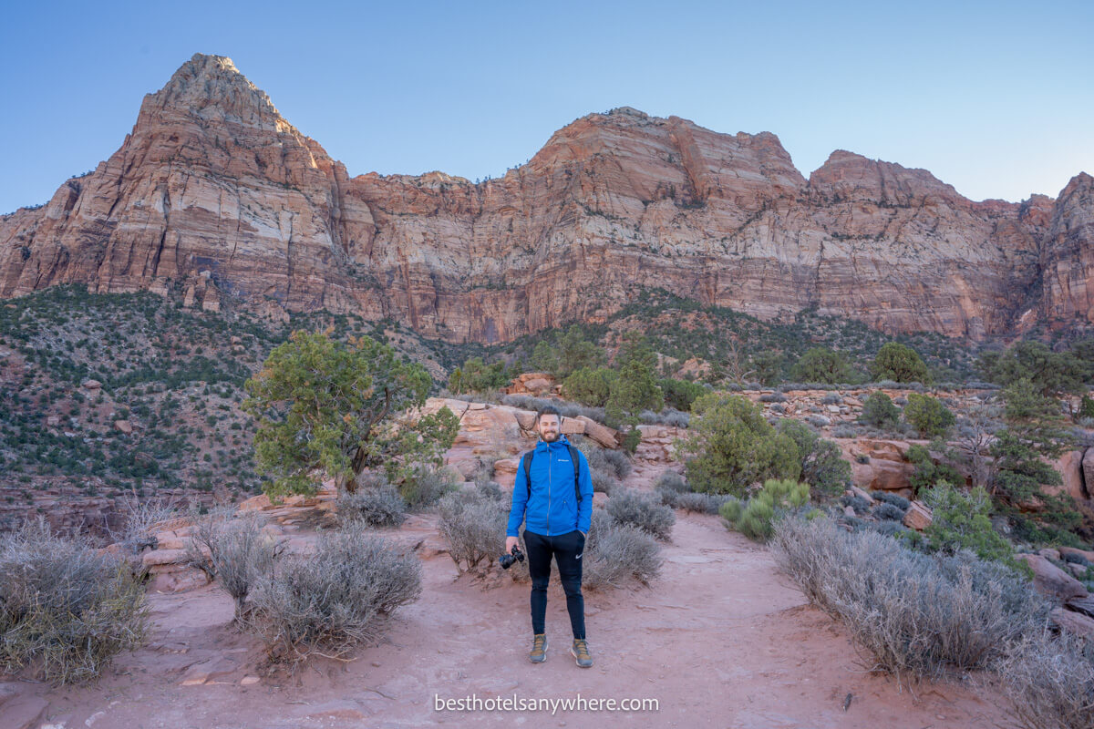 Hiker in blue coat at the summit of The Watchman Trail in Zion at dawn with blue sky