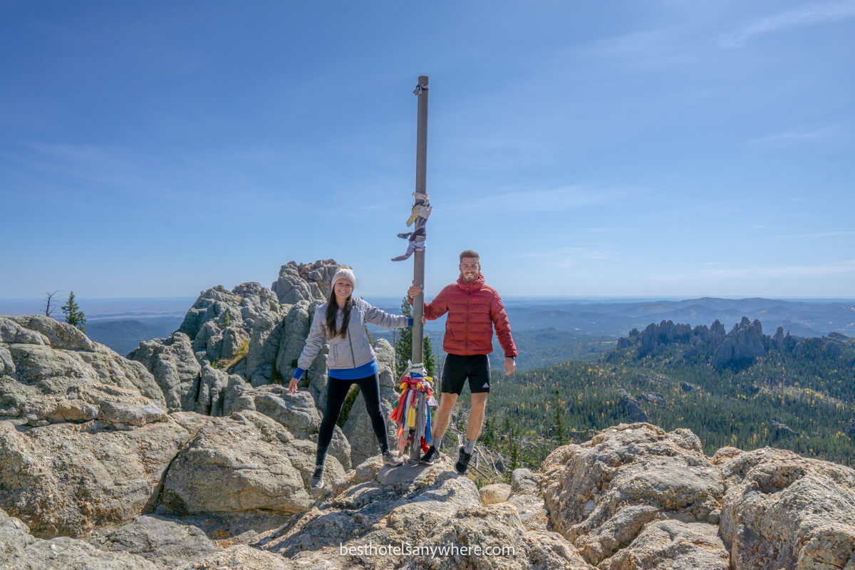 Two hikers holding onto a pole at the summit of Black Elk Peak in South Dakota on a clear day