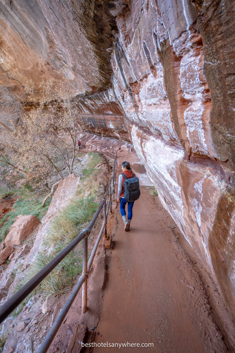 Hiker walking along a narrow path with cliff wall on one side and a rail on the other
