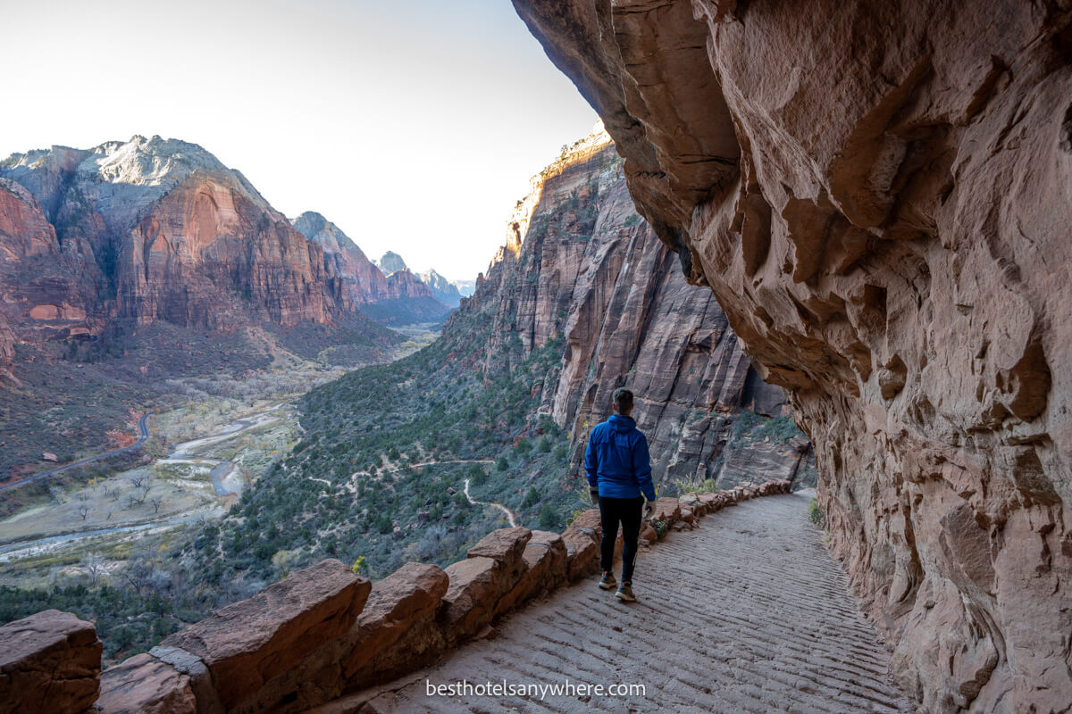 Hiker walking down a stone path with overhanging rocks and distant views of a valley and red rock cliffs