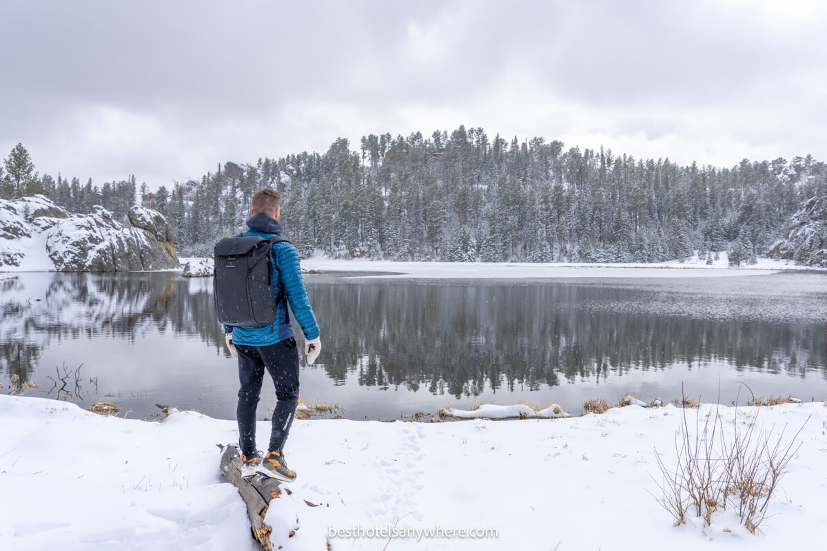 Hiker in winter gear stood on a log looking out into Sylvan Lake with snow on the ground