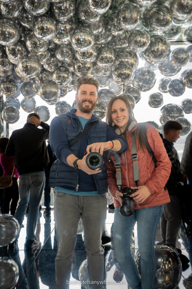 Mark and Kristen Morgan from Best Hotels Anywhere taking a photo of themselves in a mirror with silver helium balloons on the roof behind at Summit One Vanderbilt in New York City