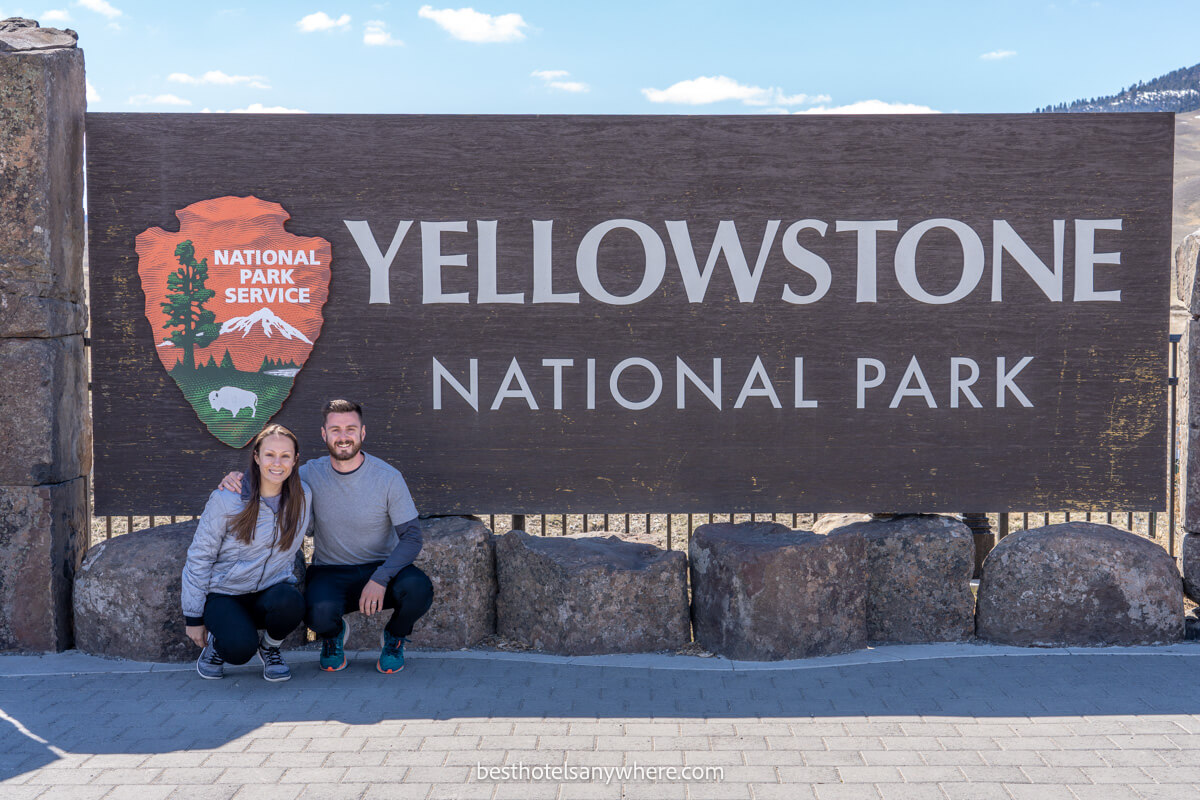 Couple crouching in the shade at Yellowstone national park entrance sign in Gardiner MT